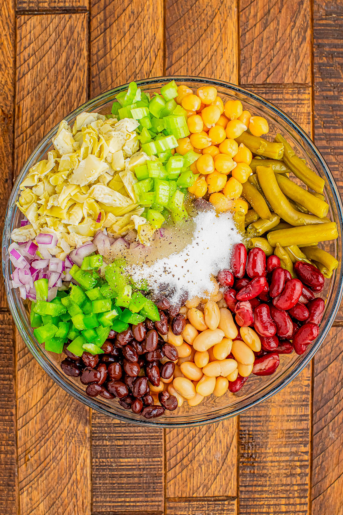 Glass bowl filled with chopped cabbage, red onion, green bell pepper, green beans, corn, kidney beans, black beans, white beans, and seasonings on a wooden surface.