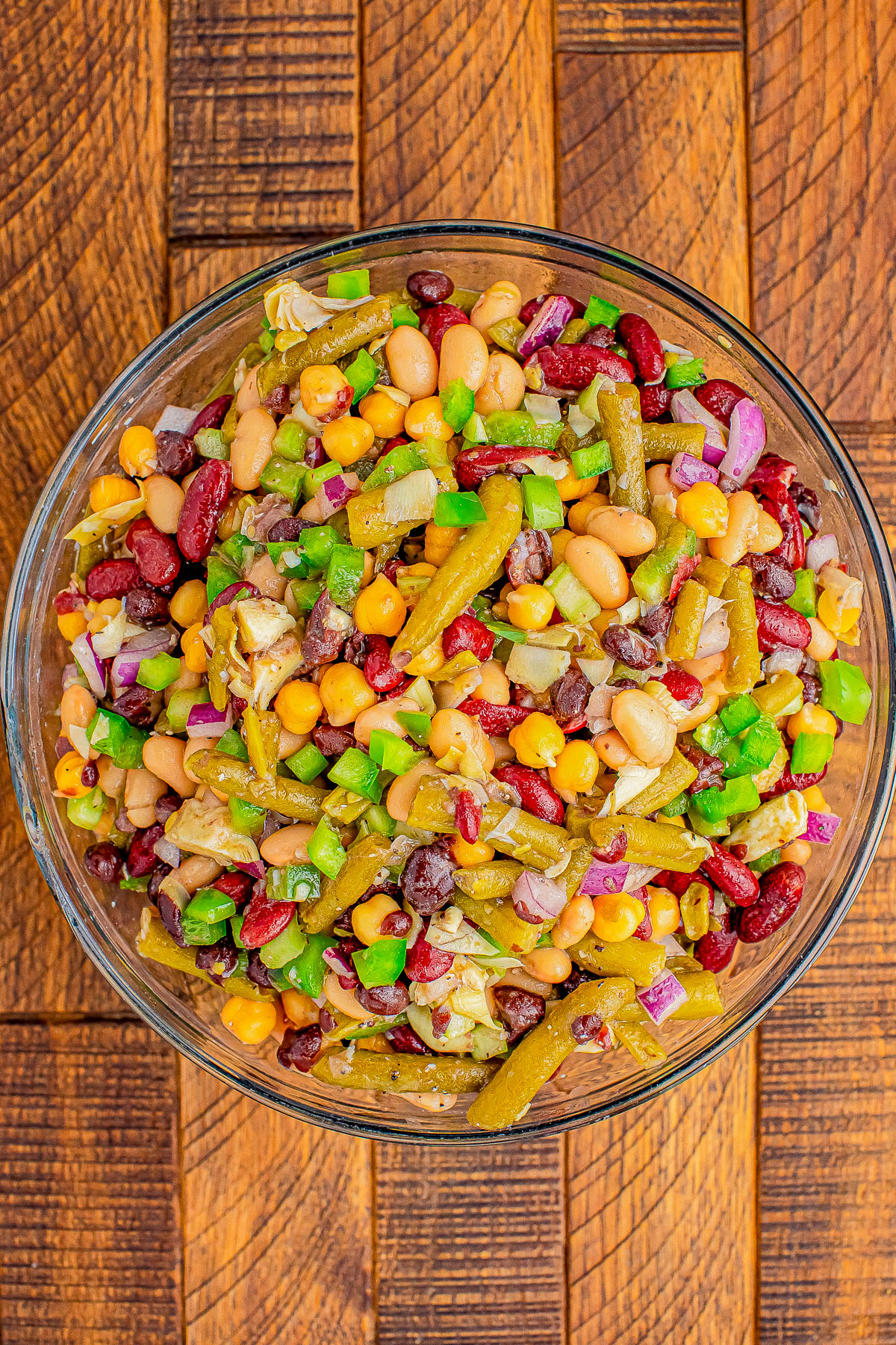 A glass bowl filled with a colorful bean salad, featuring kidney beans, white beans, green beans, corn, diced green bell peppers, and red onions, on a wooden surface.