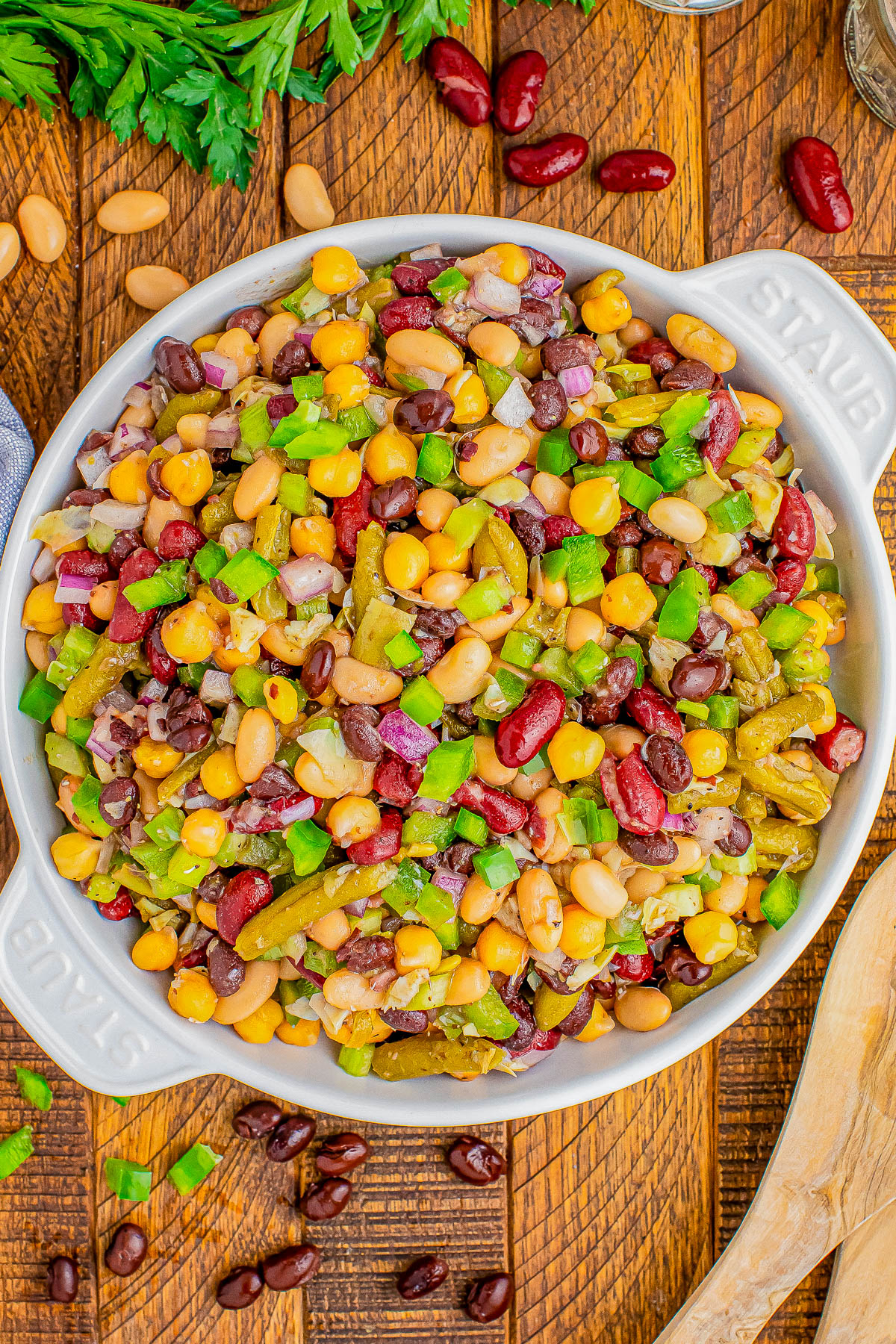 A round dish filled with a colorful bean salad, featuring red kidney beans, green bell peppers, onions, and corn kernels. Parsley and scattered beans are visible on the wooden table around the dish.