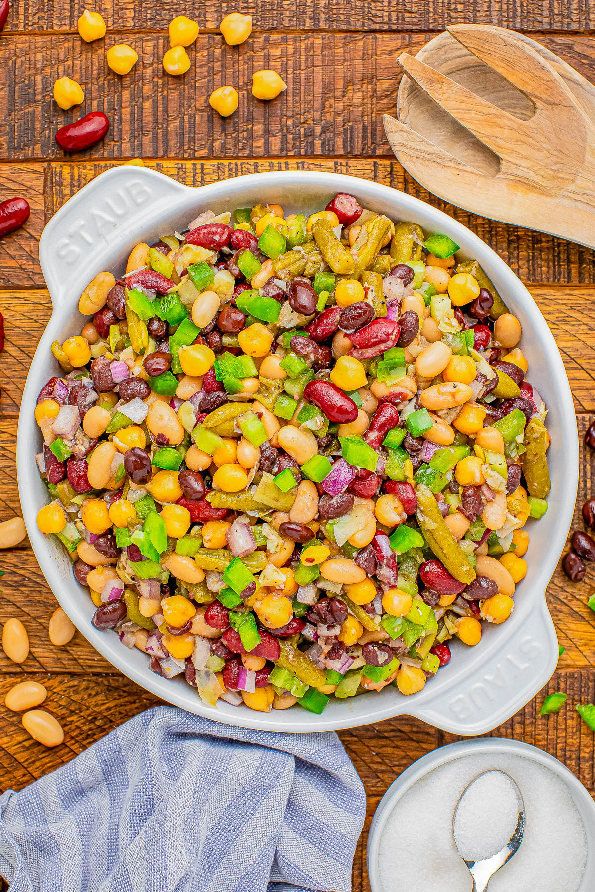 A bowl of mixed bean salad including kidney beans, chickpeas, corn, chopped green bell peppers, and onions, with a wooden spoon, a small bowl of salt, and a striped cloth nearby on a wooden surface.