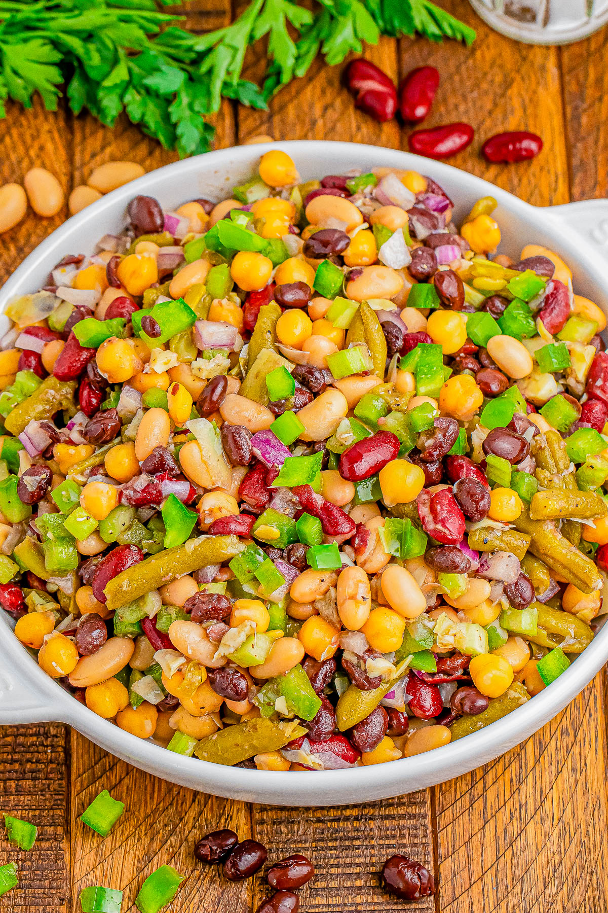 A bowl of colorful bean salad with corn, green beans, red onions, green bell peppers, and various beans, garnished with chopped parsley.