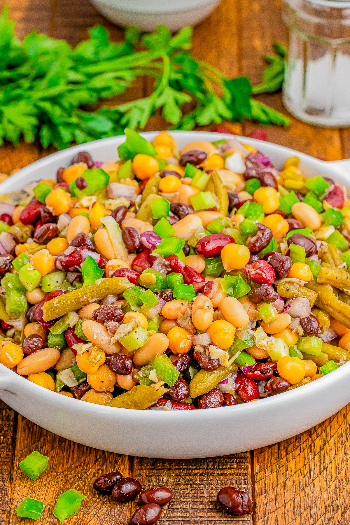 A bowl filled with a colorful mixed bean salad, featuring black beans, kidney beans, sweet corn, green peppers, and onions, garnished with parsley. A wooden table background with fresh herbs.