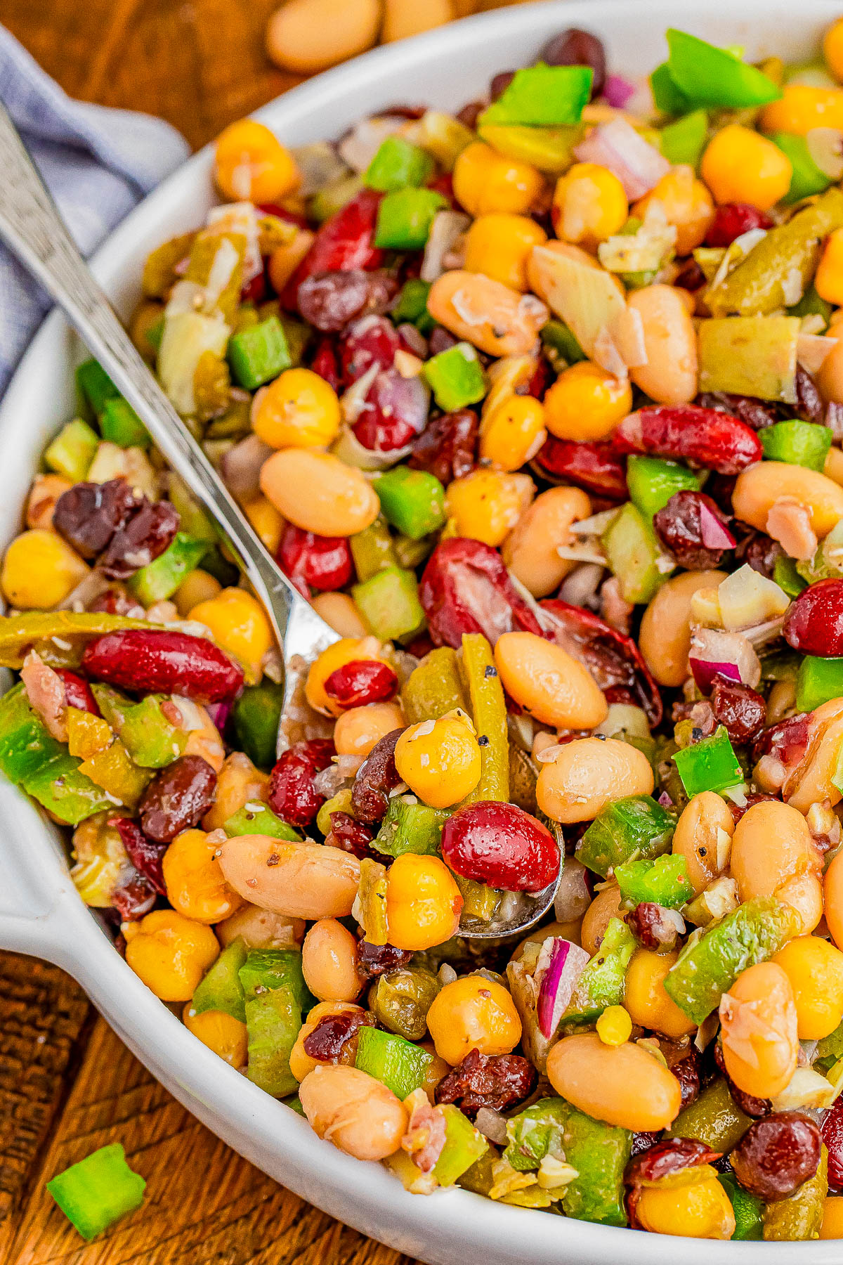 A close-up of a mixed bean salad with corn, kidney beans, black beans, green bell peppers, red onions, and a spoon in a white bowl.