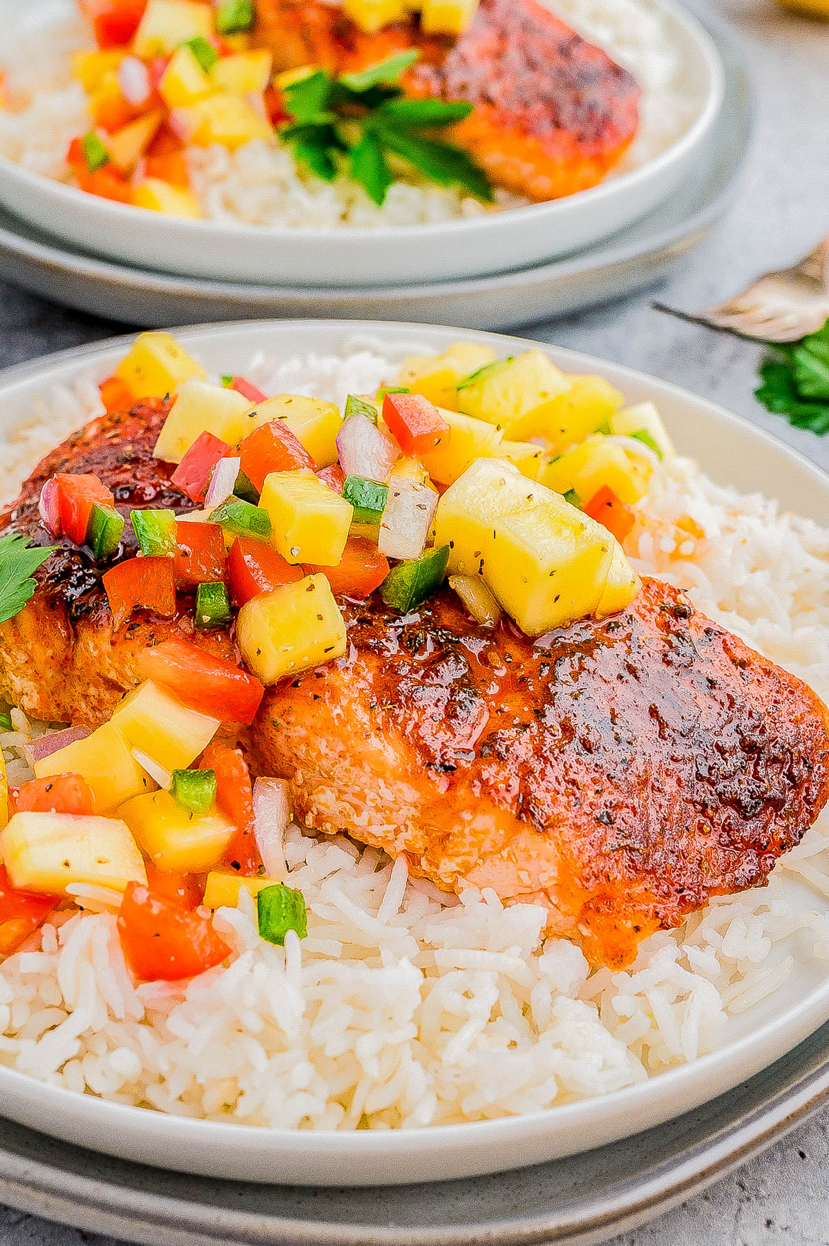 A plate of cooked white rice topped with seasoned salmon fillet and a colorful pineapple salsa. Another similar plate is in the background.