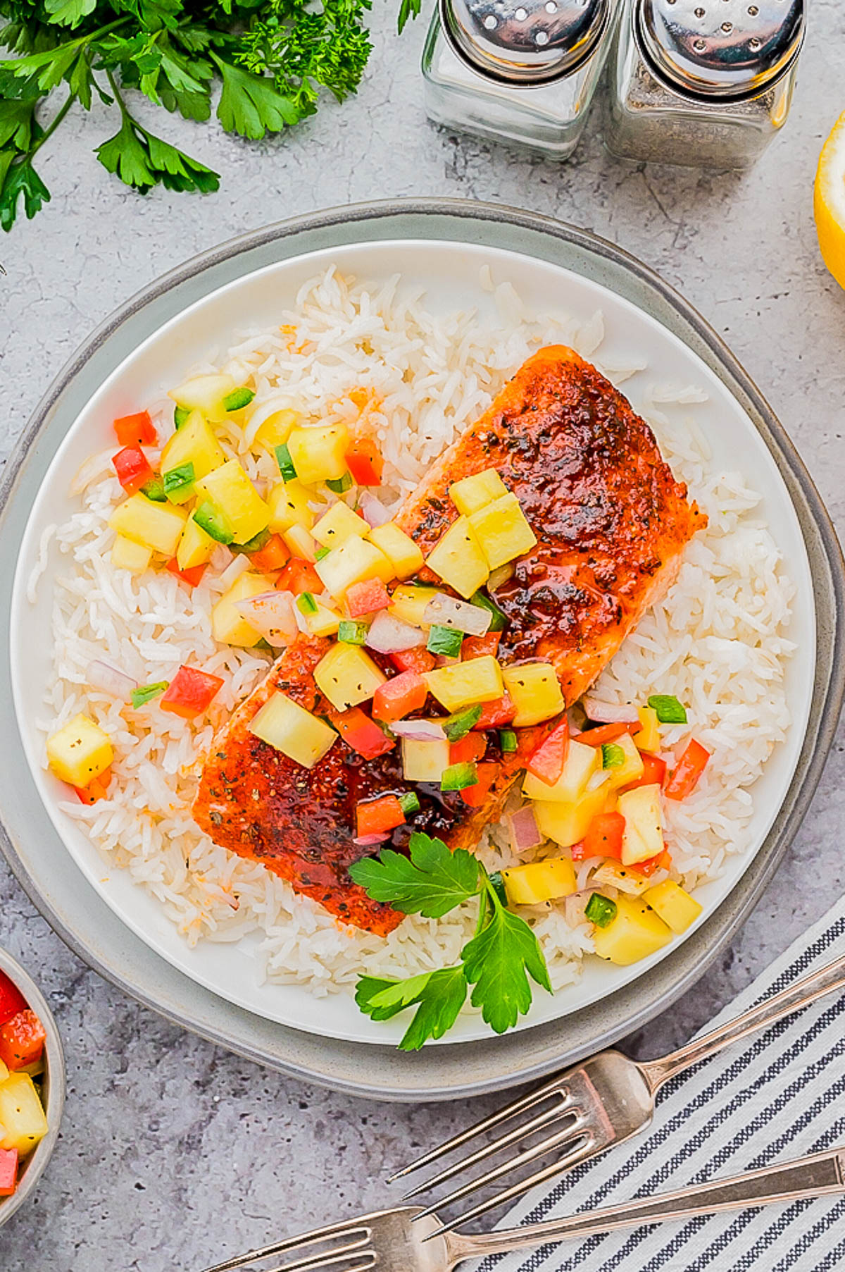 A plate of white rice topped with a grilled salmon fillet and a colorful salsa made of diced fruits and vegetables. Garnished with fresh parsley. Forks, salt, pepper shakers, and parsley are nearby.