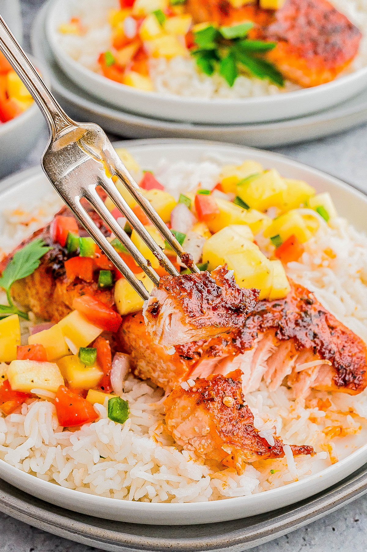 A fork holding a piece of grilled salmon over a bed of rice, garnished with diced mango, tomatoes, and herbs.