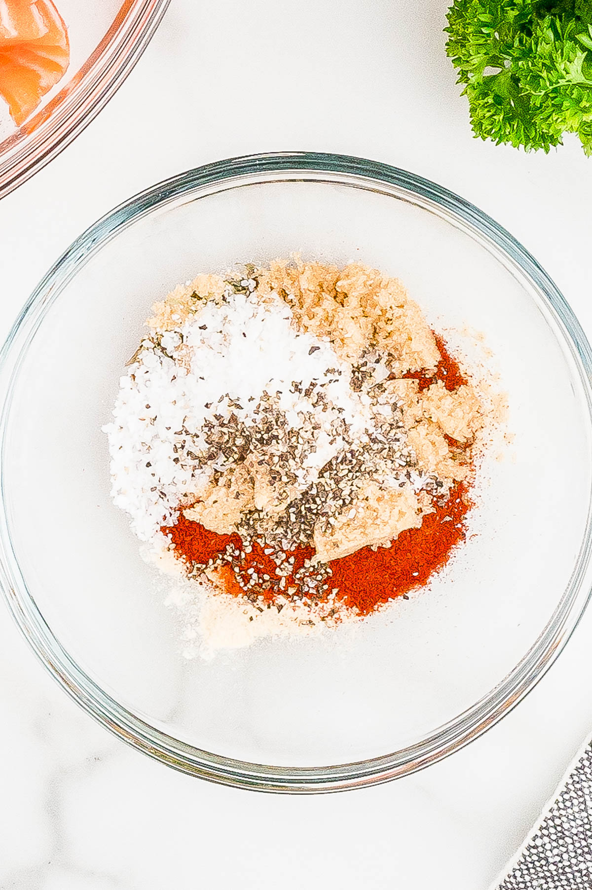 A glass bowl containing a mixture of spices, including salt, black pepper, garlic powder, onion powder, and paprika, with a bunch of parsley in the background.