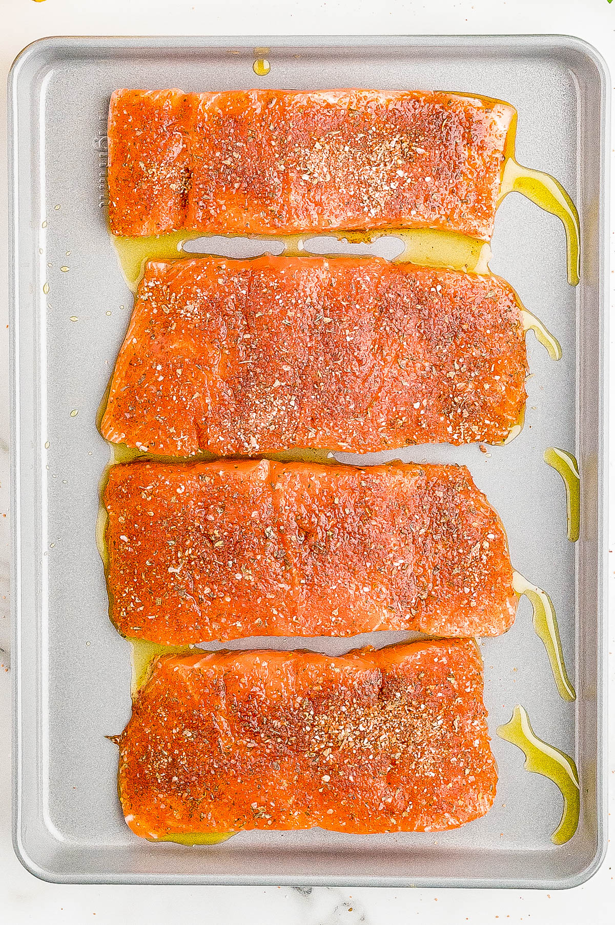 Four seasoned salmon fillets are arranged on a baking sheet, drizzled with olive oil, prepared for cooking.