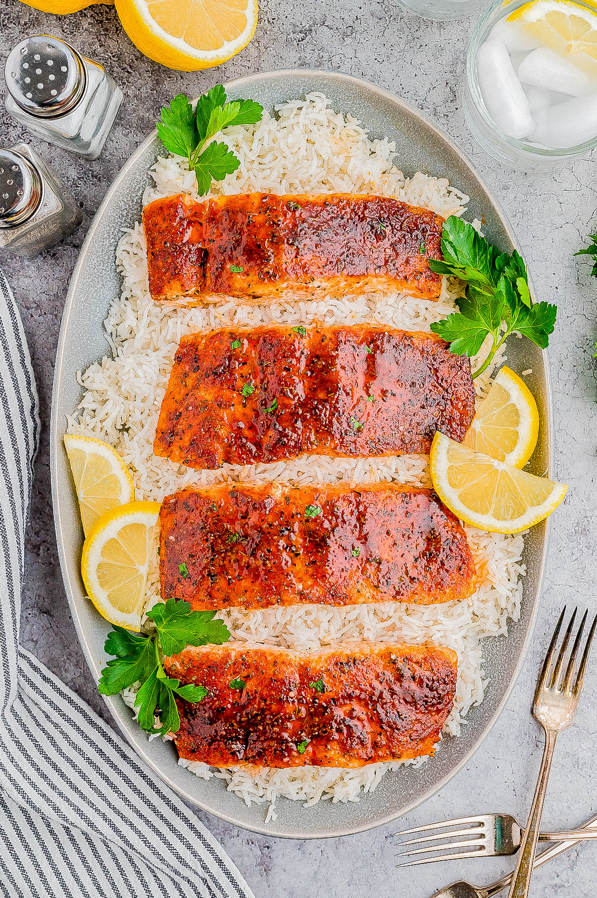 A dish of four seasoned salmon fillets on a bed of white rice, garnished with parsley and lemon slices. Silverware, salt and pepper shakers, and a glass of water with ice are nearby.