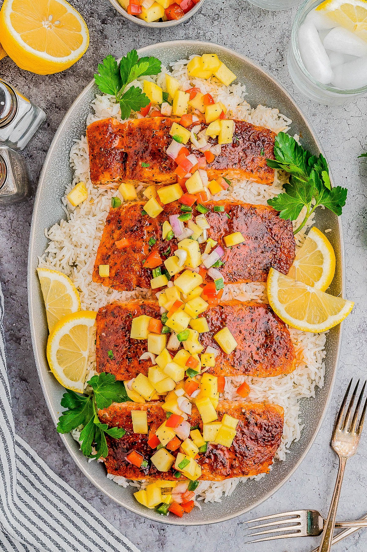 A plate of rice topped with four fillets of seasoned grilled fish, garnished with a chunky pineapple salsa, lemon slices, and parsley. A lemon half, fork, and striped napkin are also visible.