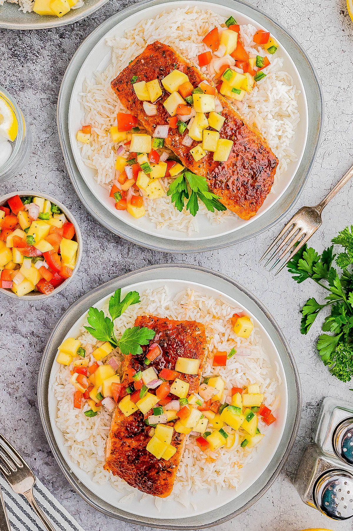 Two plates of grilled salmon topped with fruit salsa served over white rice, with garnishes and additional salsa on the side. A fork and various green herbs surround the plates.