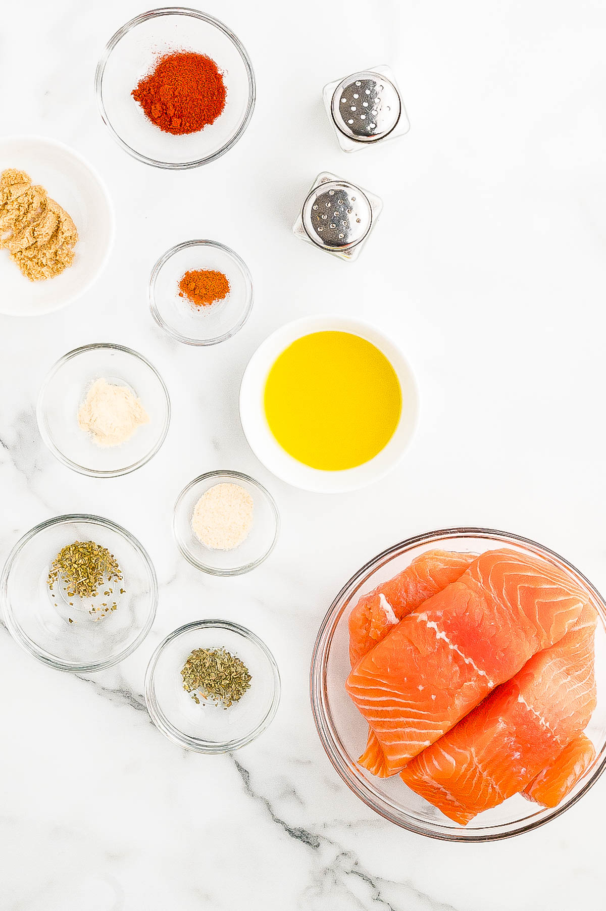 Salmon fillets in a bowl with various spices and seasonings in separate small bowls, including paprika, brown sugar, garlic powder, dried herbs, and a dish of olive oil, on a white marble surface.