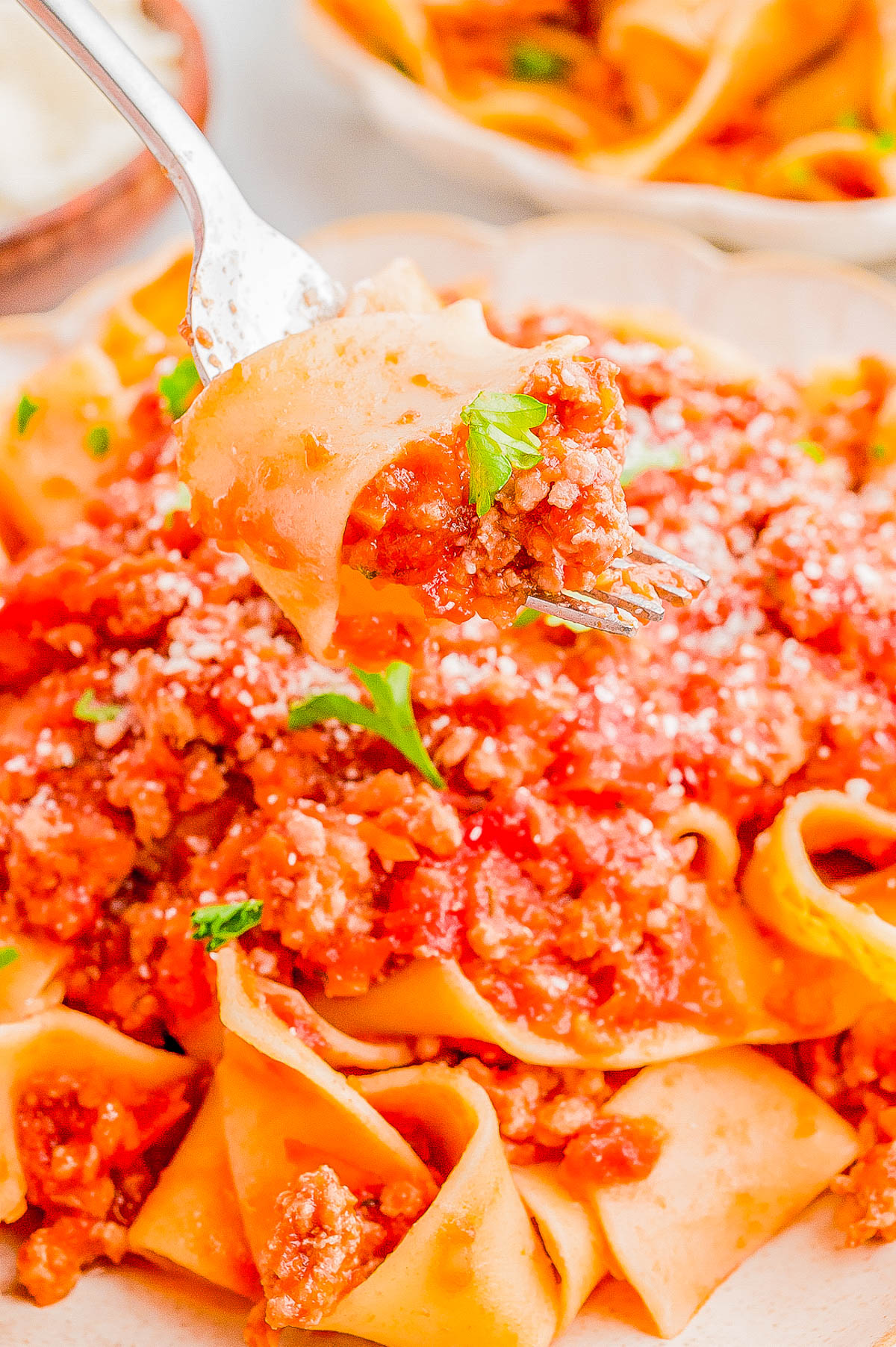 A fork lifts a bite of pappardelle pasta with a chunky meat and tomato sauce garnished with parsley from a plate of the same pasta dish.