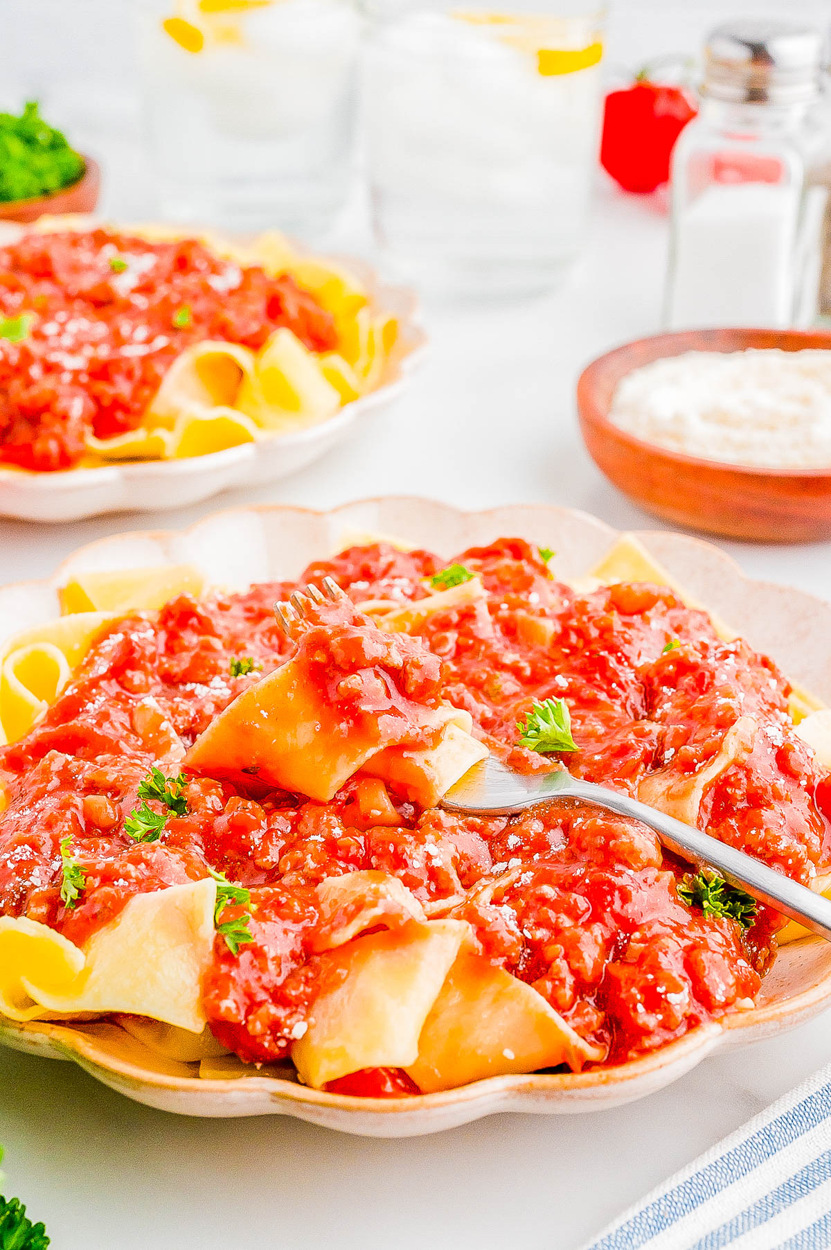 A plate of pasta topped with a red tomato-based sauce, garnished with parsley, is shown with a fork. Two glasses with ice and lemon slices, and a bowl of grated cheese are in the background.