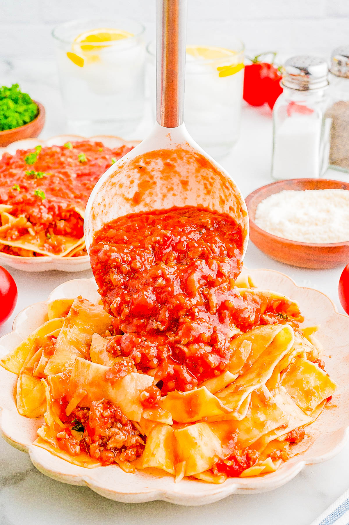 A ladle pours a chunky tomato meat sauce over a plate of pasta. Shredded cheese, salt and pepper shakers, lemon water, and fresh parsley are in the background.