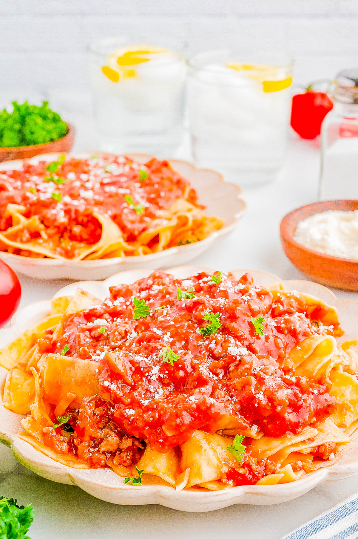 Two plates of pasta topped with a generous amount of tomato-based sauce and garnished with parsley, accompanied by two glasses of water with lemon slices, a bowl of grated cheese, and fresh herbs on the table.