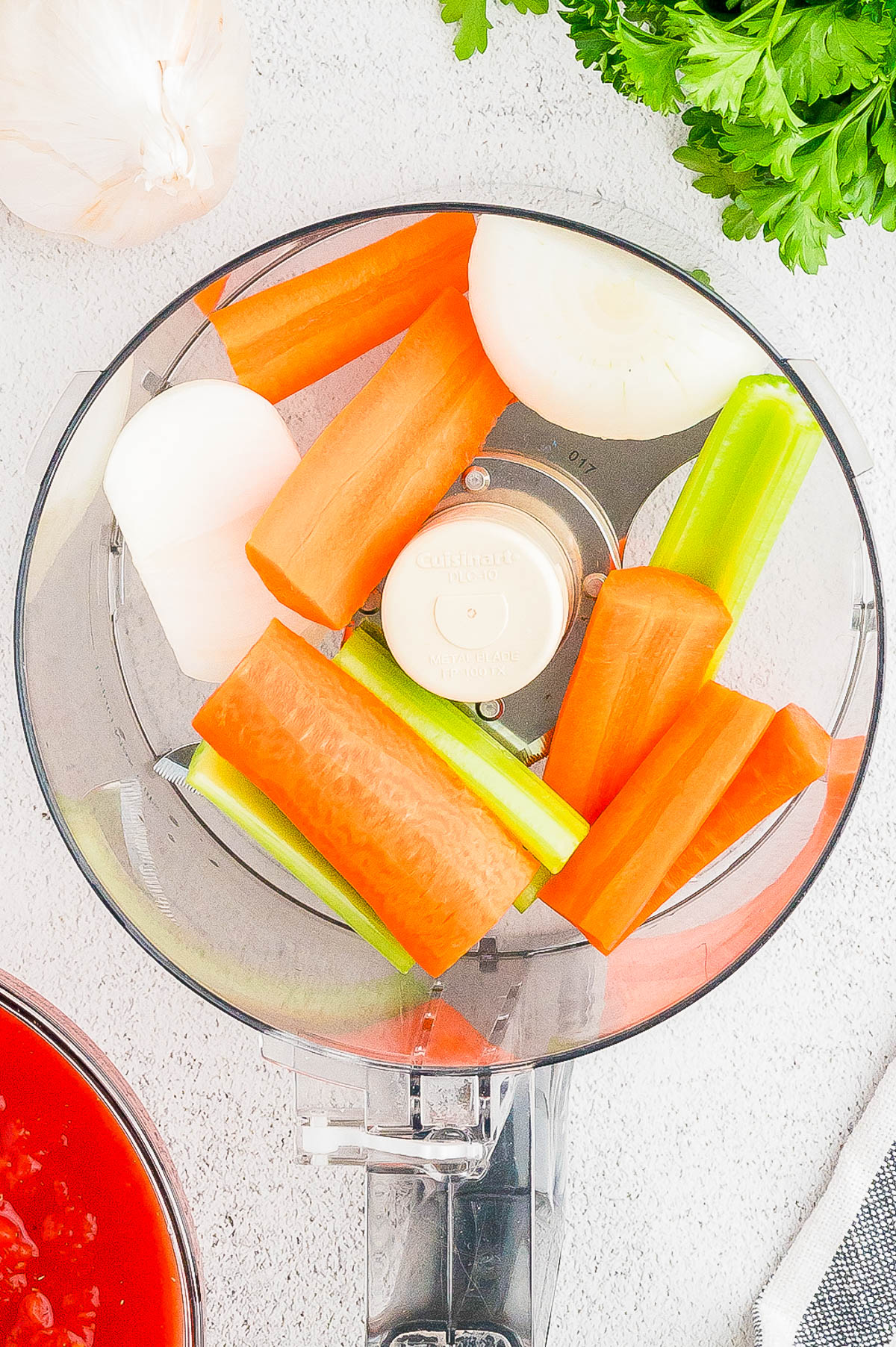 Top view of a food processor containing roughly chopped carrots, onions, and celery. Partial views of a garlic bulb, parsley, a red liquid in a bowl, and a striped cloth are visible in the surrounding area.