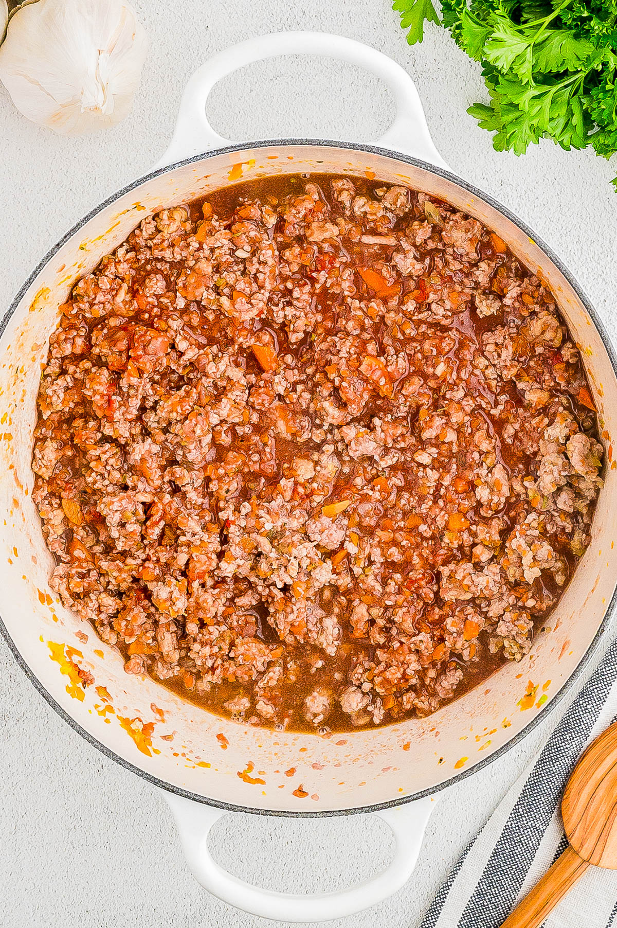 A white pot containing a cooked ground meat mixture with a tomato-based sauce on a light-colored surface, garnished with garlic cloves and fresh herbs nearby.