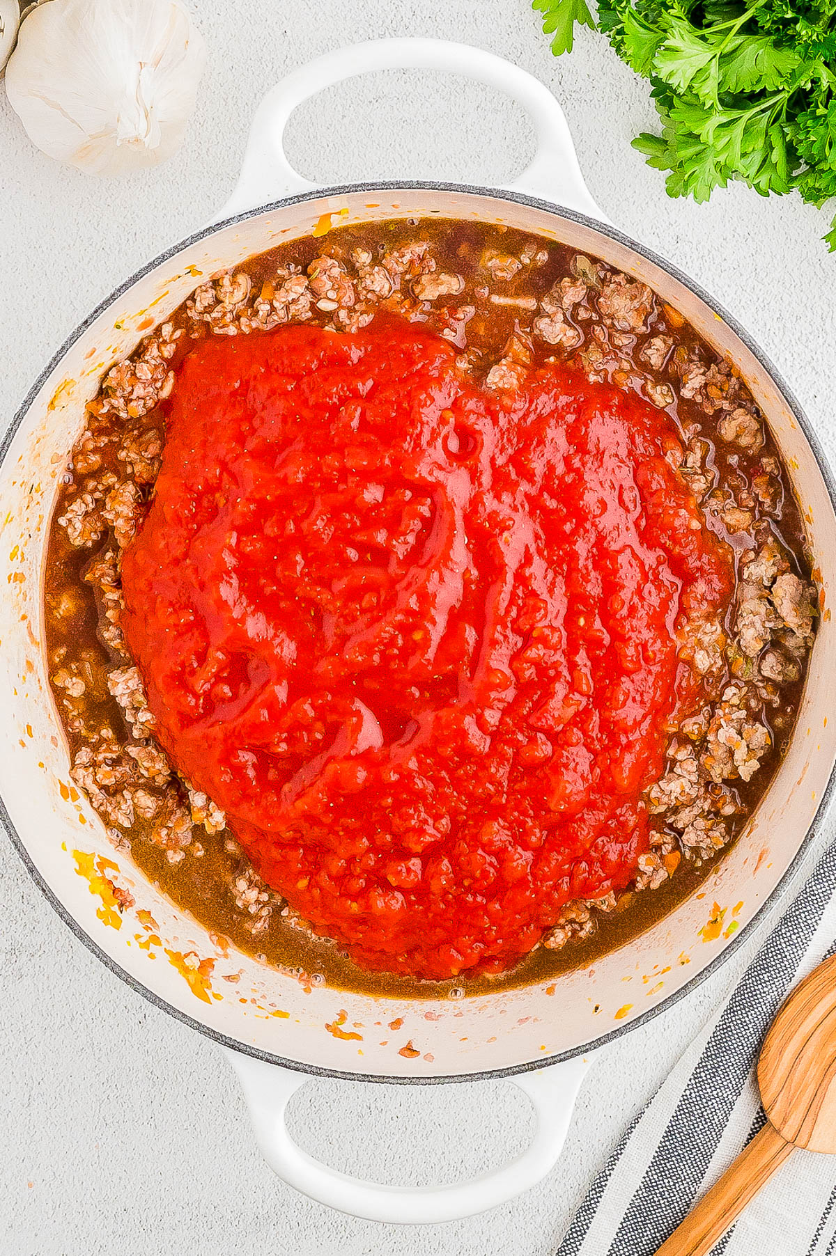 A pot of ground meat and tomato sauce cooking on a stove, surrounded by garlic, fresh parsley, and wooden utensils.