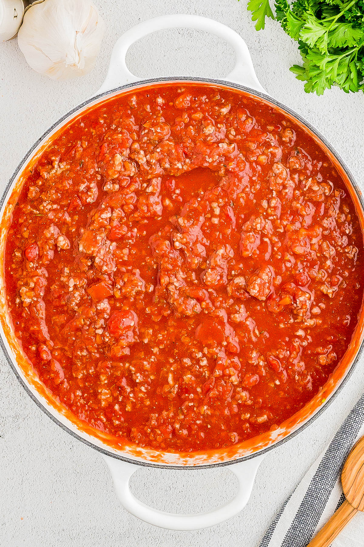 A pot filled with tomato-based meat sauce, placed on a light-colored surface next to a garlic bulb, parsley, and a wooden spoon.