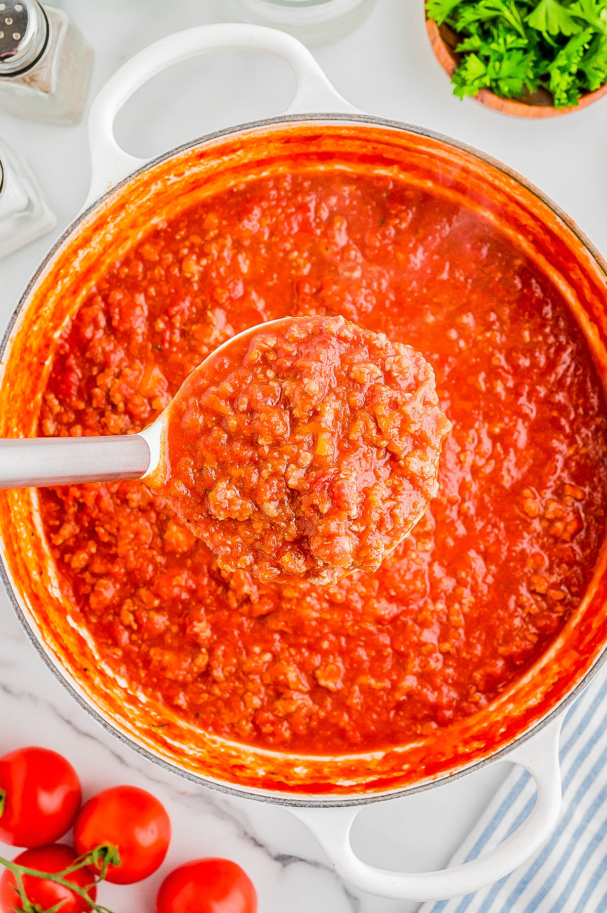 A ladle filled with tomato-based meat sauce is lifted from a pot. The surrounding area includes fresh tomatoes, parsley, salt, and pepper shakers on a white surface.