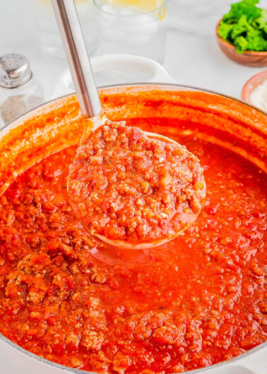 A ladle filled with hearty tomato and meat sauce hovers above a pot of the same sauce. Ingredients like parsley and salt are blurred in the background.