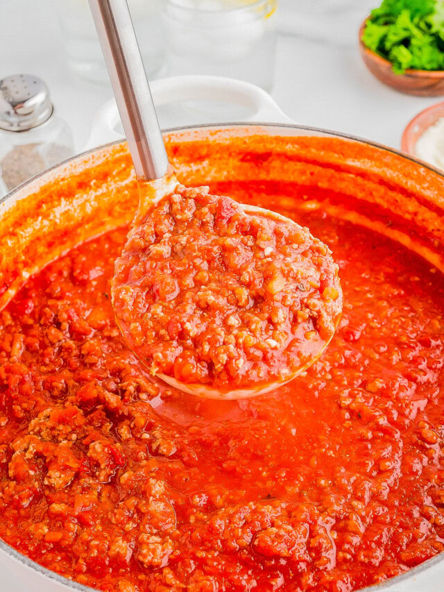 A ladle filled with hearty tomato and meat sauce hovers above a pot of the same sauce. Ingredients like parsley and salt are blurred in the background.