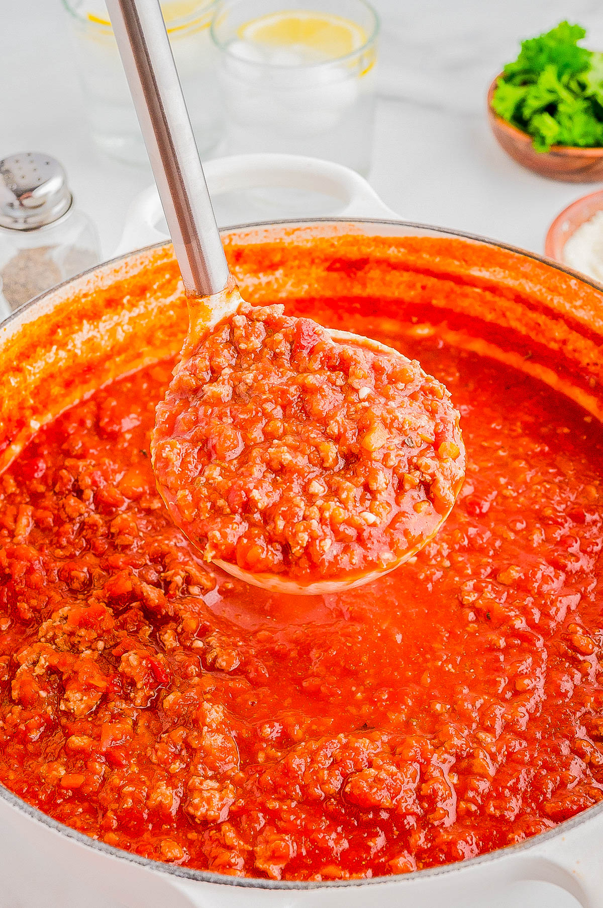 A ladle filled with hearty tomato and meat sauce hovers above a pot of the same sauce. Ingredients like parsley and salt are blurred in the background.