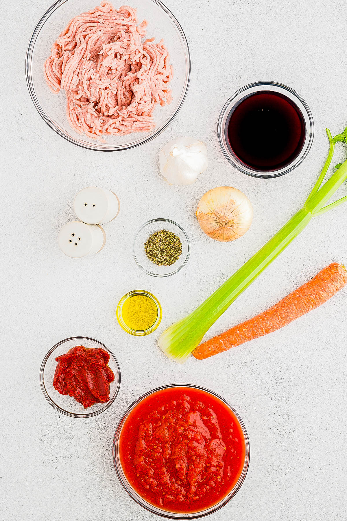 Top view of minced meat, red wine, tomato paste, crushed tomatoes, olive oil, carrot, celery, onion, garlic, salt and pepper shakers, and dried herbs on a white surface.