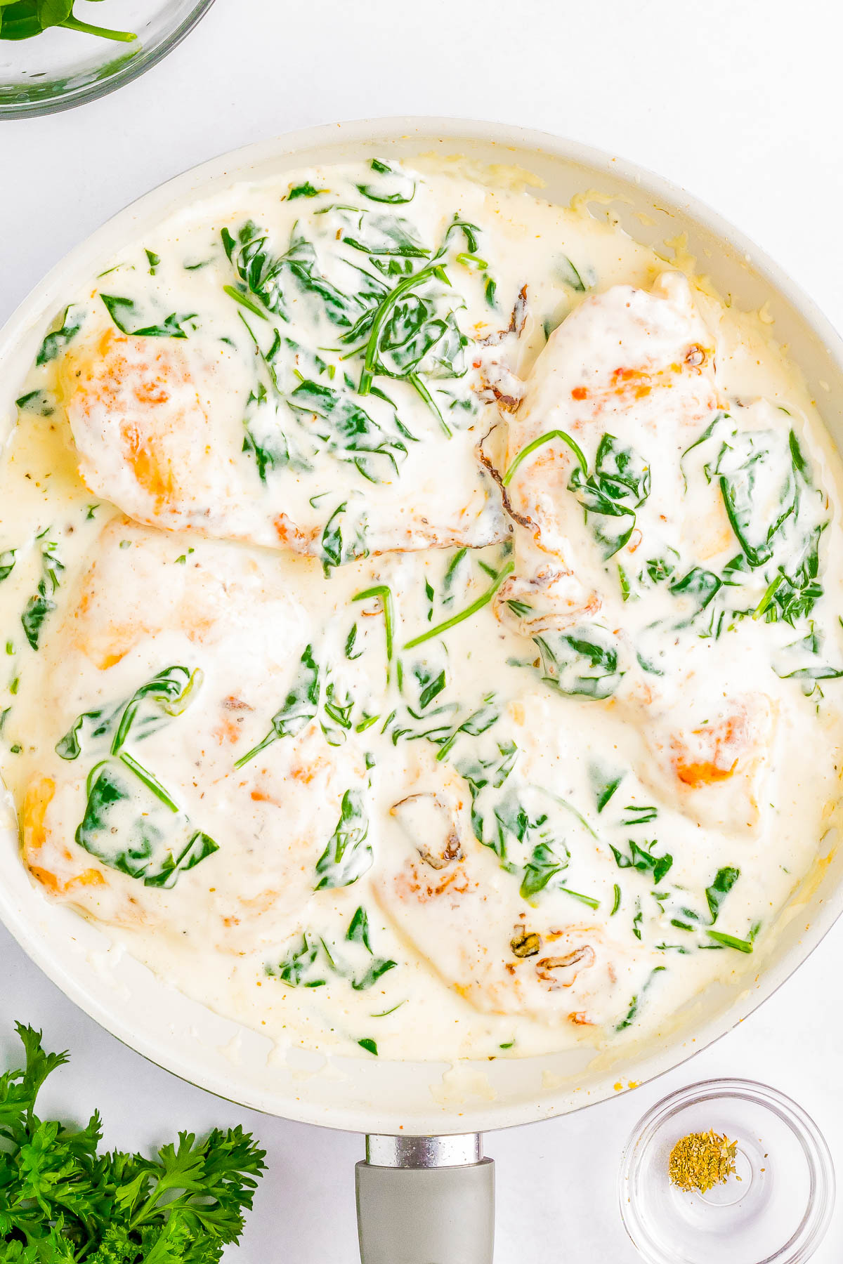 A skillet containing cooked chicken breasts topped with a creamy spinach sauce, accompanied by small bowls of spices and herbs on the side.