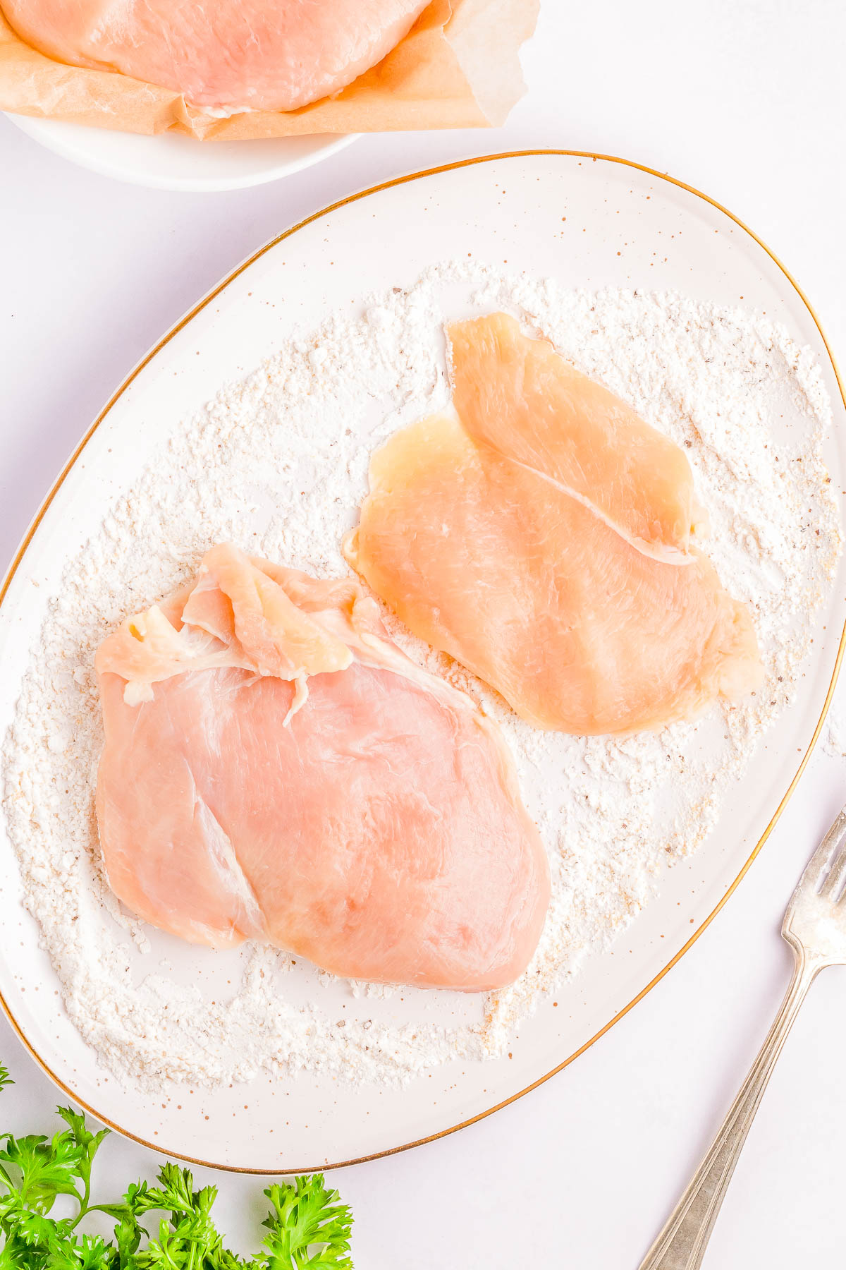 Two raw chicken breasts placed on a white plate with flour, accompanied by a fork and parsley in the corner.