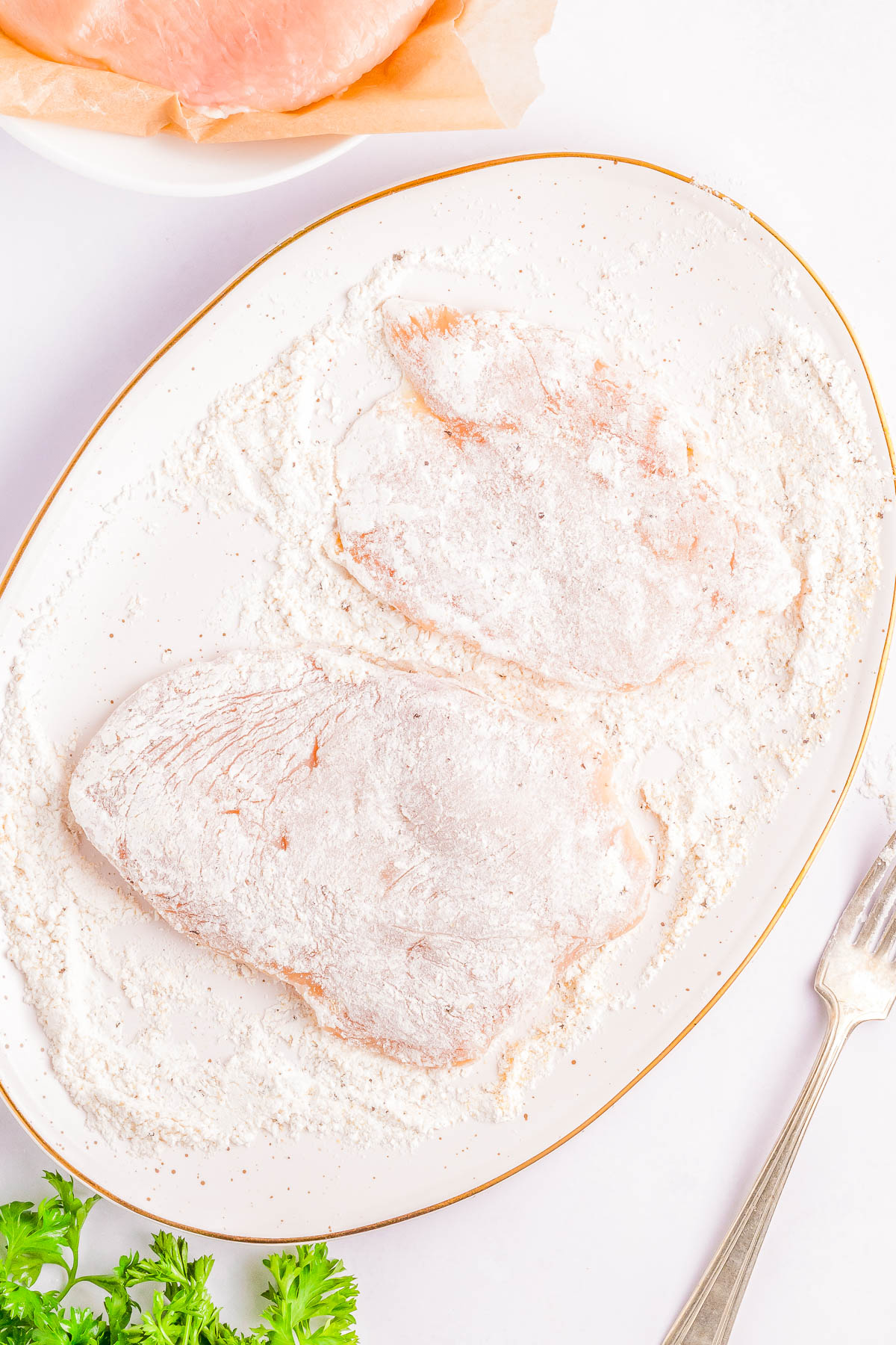 Two raw chicken breasts coated in flour lie on a white plate, with a fork and sprigs of parsley next to the plate. Another piece of chicken is partially visible in the background.