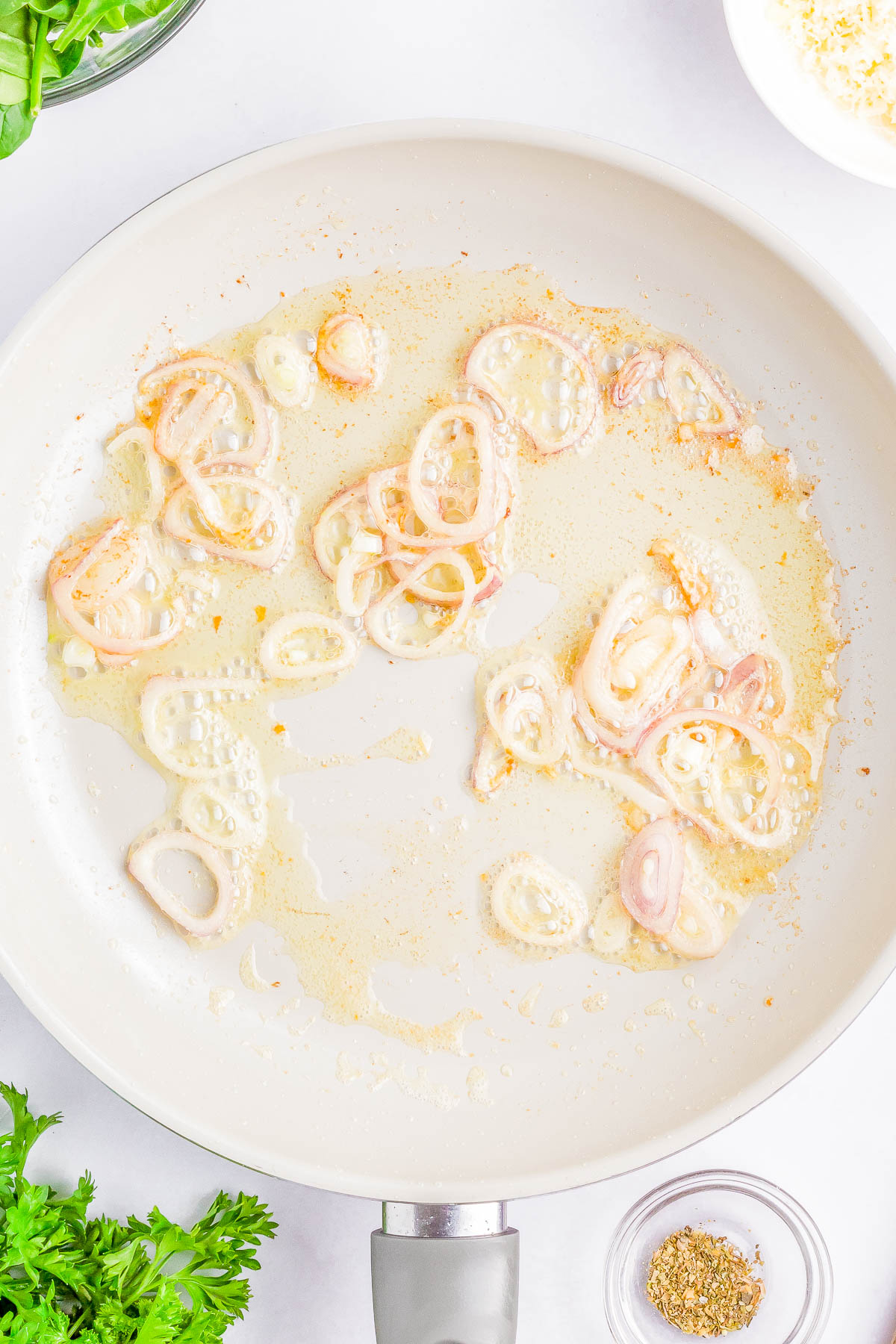 Sliced shallots sautéing in oil in a white pan, surrounded by herbs and spices on a white surface.