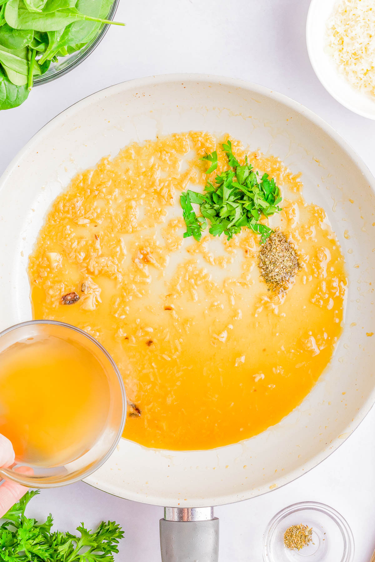 Cooking process in a pan with sautéed garlic, fresh herbs, and spices, while a hand pours broth into the mixture. Various ingredients surround the pan.