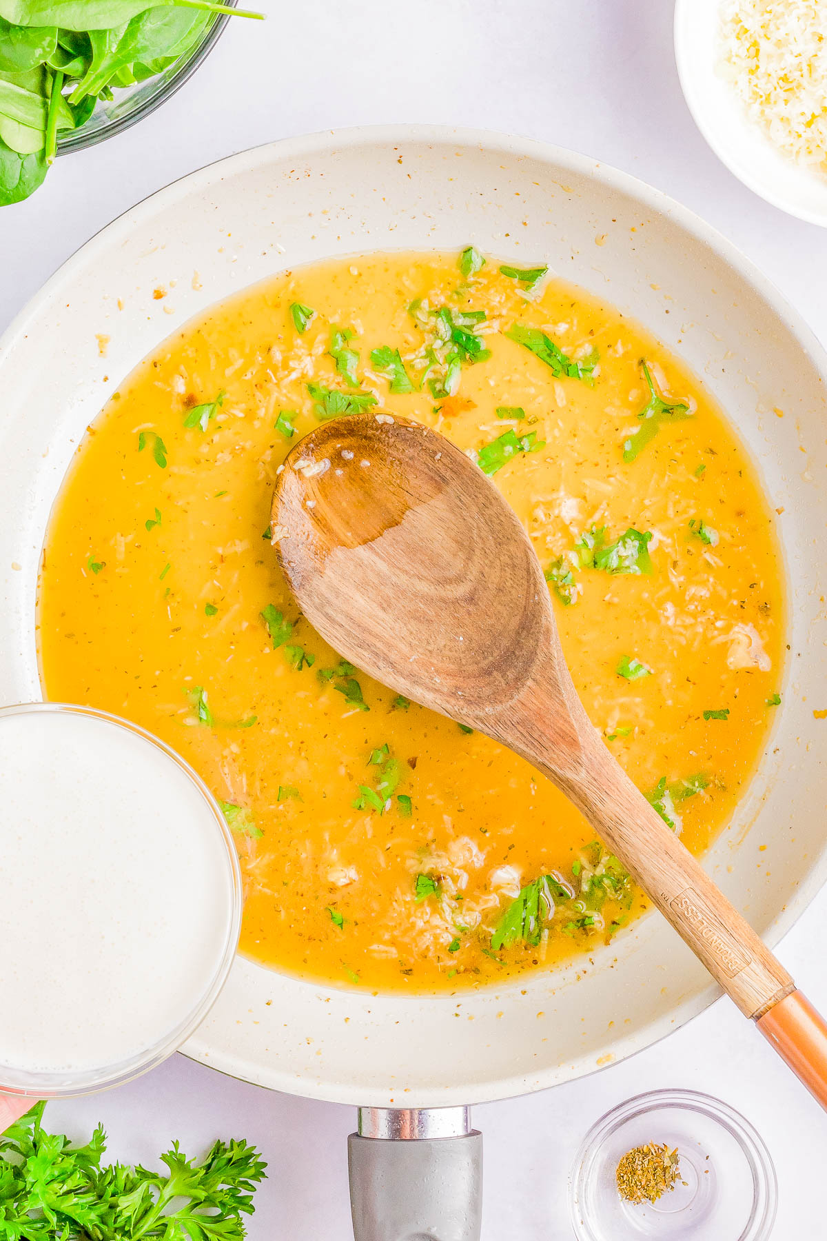 A wooden spoon rests in a skillet filled with a yellow sauce mixed with herbs. A person's hand holds a small bowl of cream over the skillet, and ingredients like greens and seasonings surround the scene.