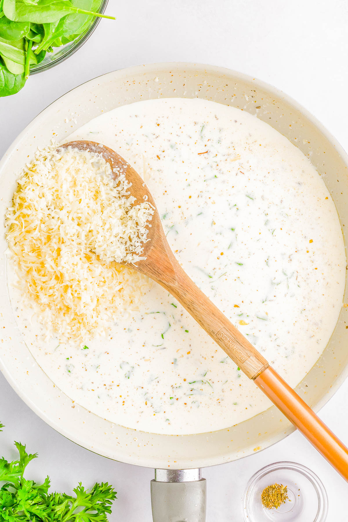 A wooden spoon stirring grated cheese into a creamy white sauce in a pan, surrounded by fresh green herbs and a small bowl of seasoning.