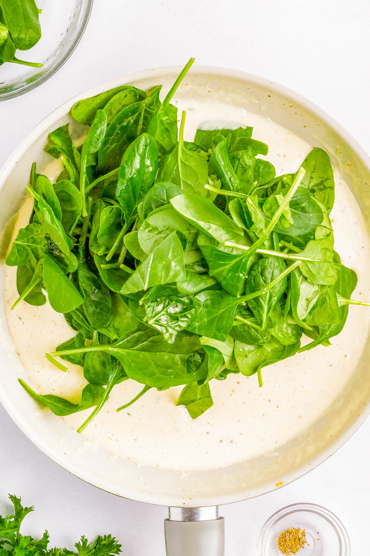 A skillet filled with a creamy sauce and topped with fresh spinach leaves sits on a light-colored surface. Surrounding the skillet are small bowls containing spices and herbs.