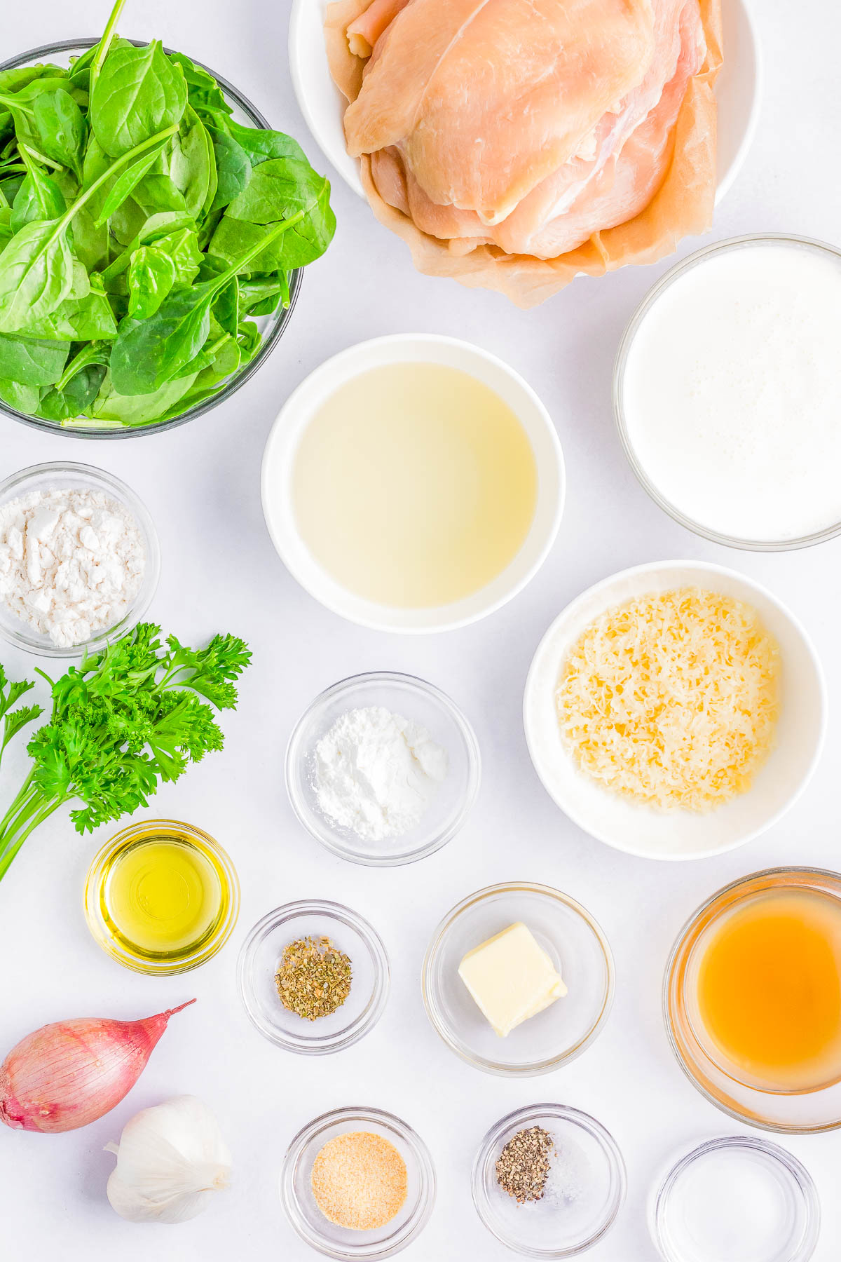 An array of ingredients including raw chicken, spinach, parsley, a shallot, garlic, assorted spices, flour, oil, butter, chicken broth, cream, and grated cheese arranged on a white surface.