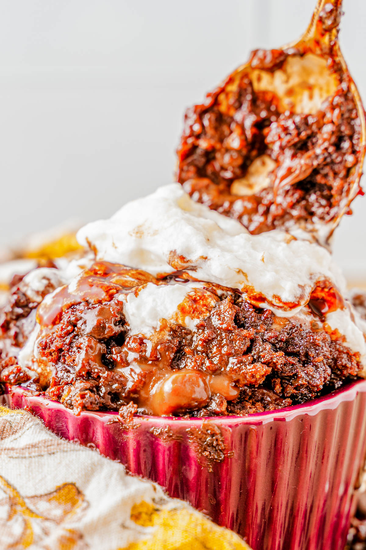 Close-up of a rich, chocolate lava cake in a red ramekin, topped with a generous serving of melted marshmallow and a spoon scooping out the gooey center.