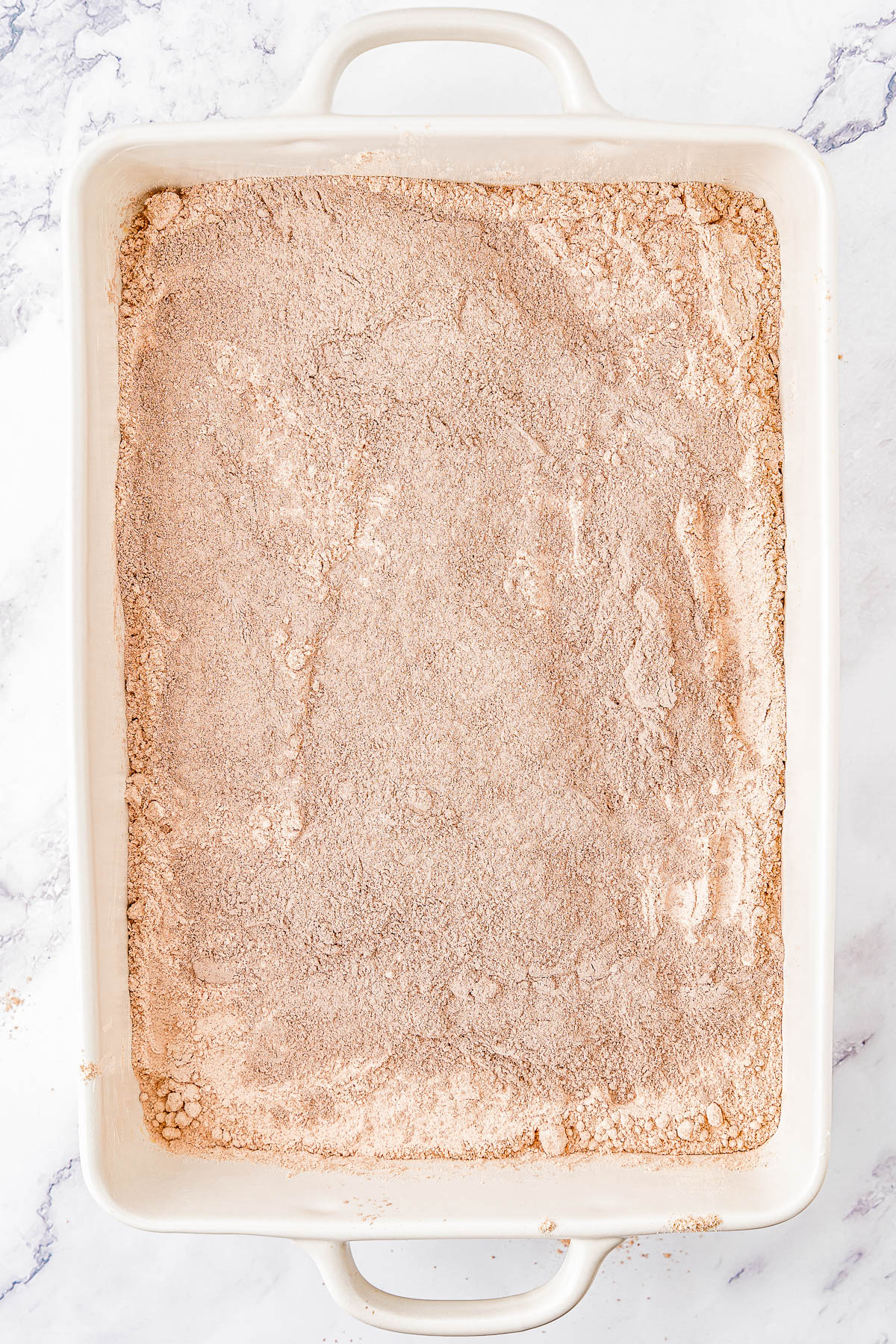 A rectangular baking dish filled with a layer of dry, brown-colored mixture, likely cake or bread mix, placed on a marble surface.