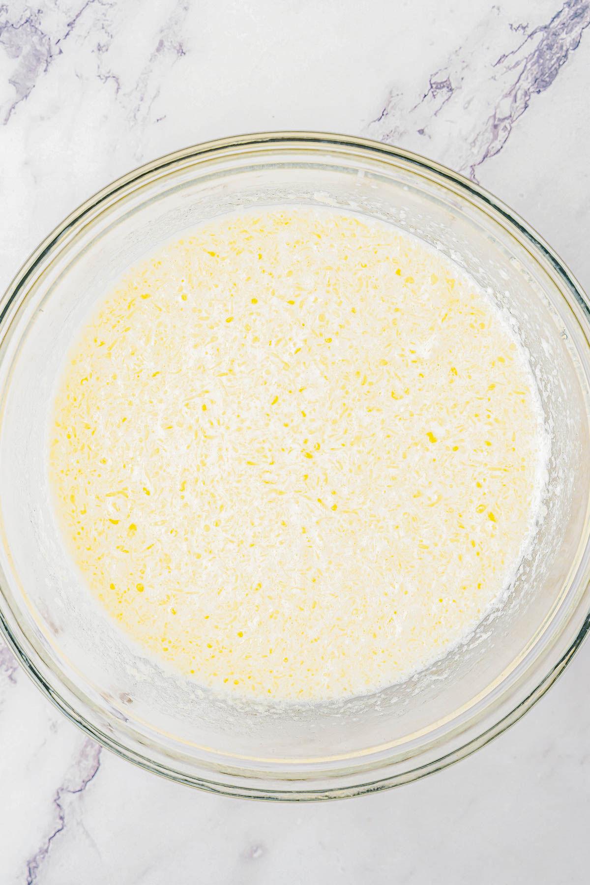 A large glass bowl containing a mixture of dry and wet ingredients for corn cake batter, set on a marbled surface.