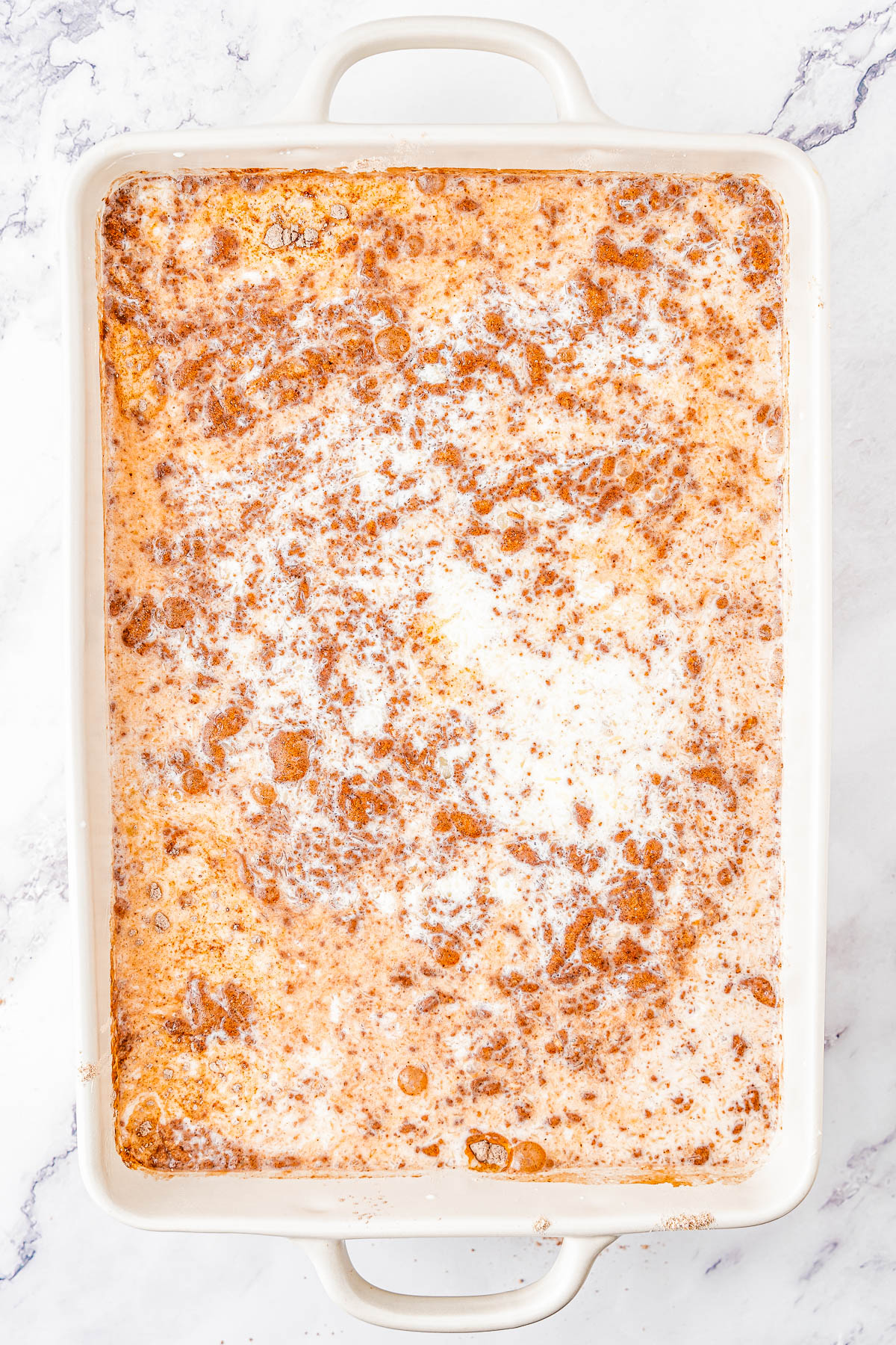 A rectangular white casserole dish filled with a baked mixture, showing a golden-brown crust with a marbled appearance of cream and cinnamon. The dish is placed on a white marble surface.