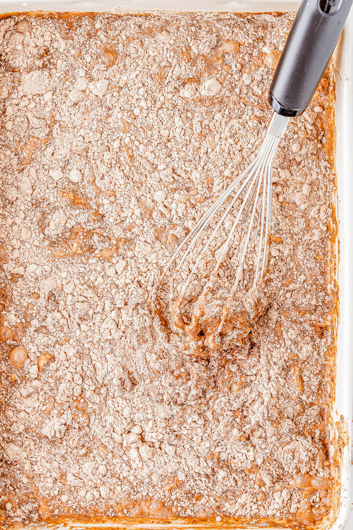 A whisk is seen partially submerged in a rectangular baking dish filled with a mixture of batter and a crumbly topping.