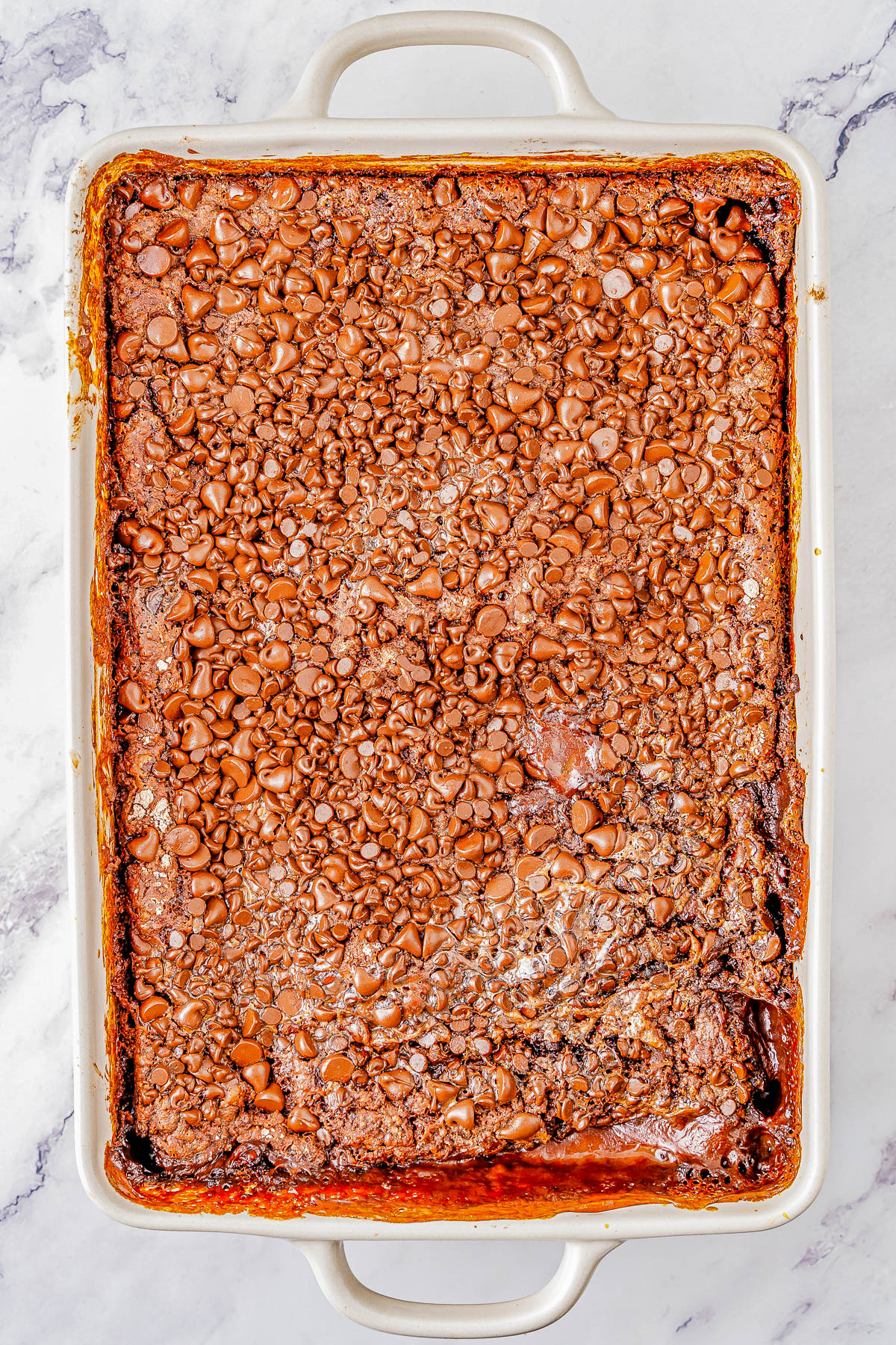 A baked dessert in a rectangular dish topped with melted chocolate chips, viewed from above on a marble surface.