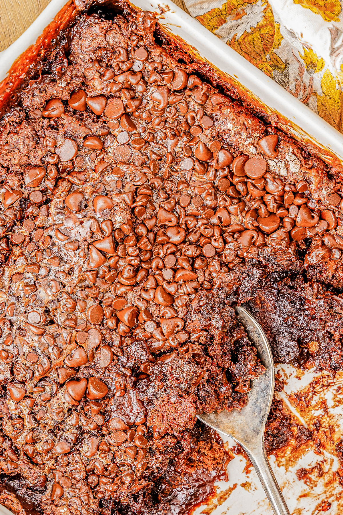 A brownie dessert with a melted chocolate topping is shown in a rectangular baking dish. A spoon is scooping out a portion from the corner.