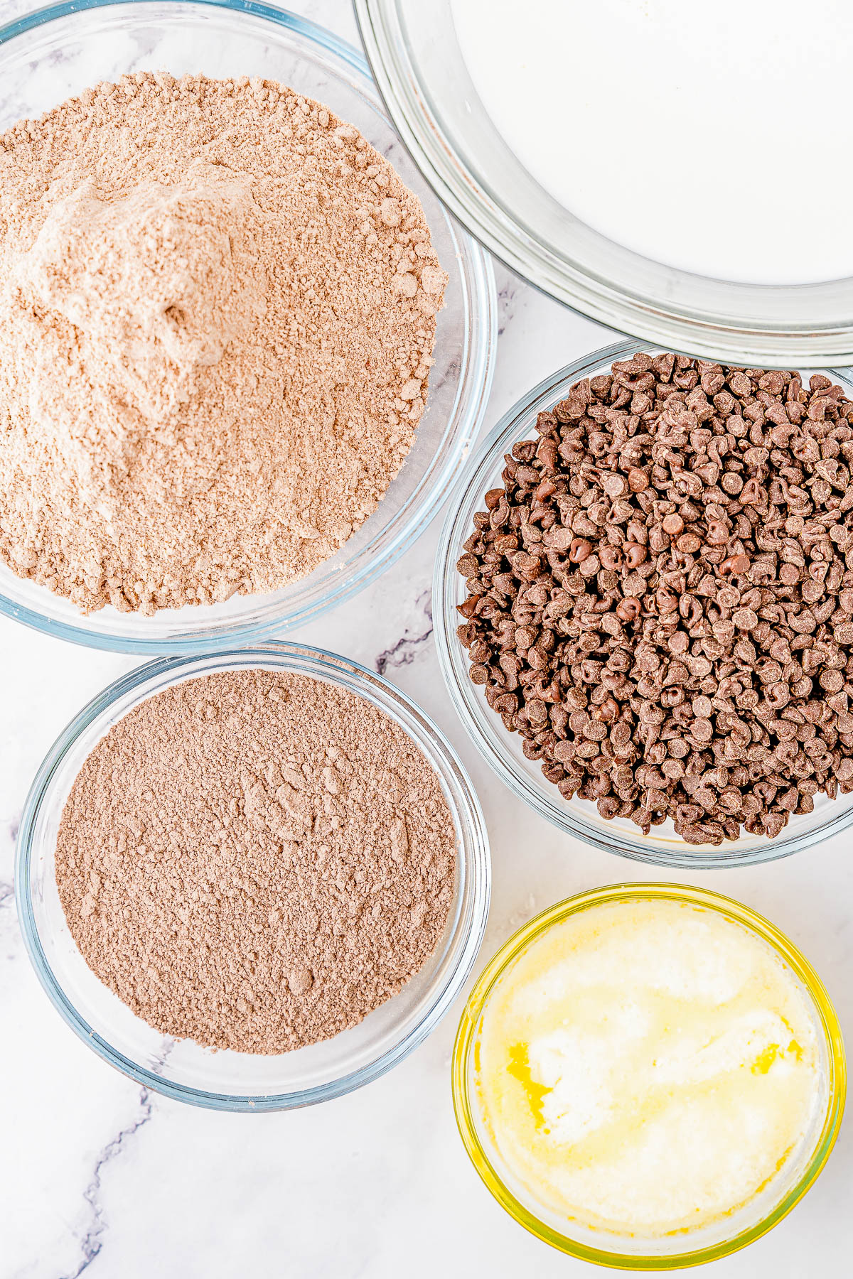 Overhead view of five bowls containing flour, chocolate chips, cocoa powder, melted butter, and milk, arranged on a marble surface.