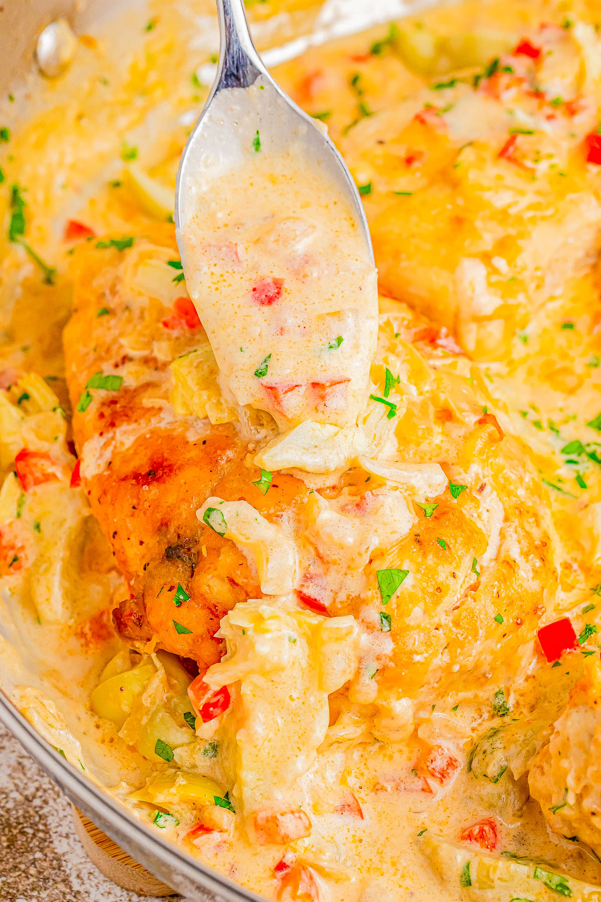 A spoon drizzles creamy sauce over chicken breasts cooked with diced peppers and herbs in a skillet.