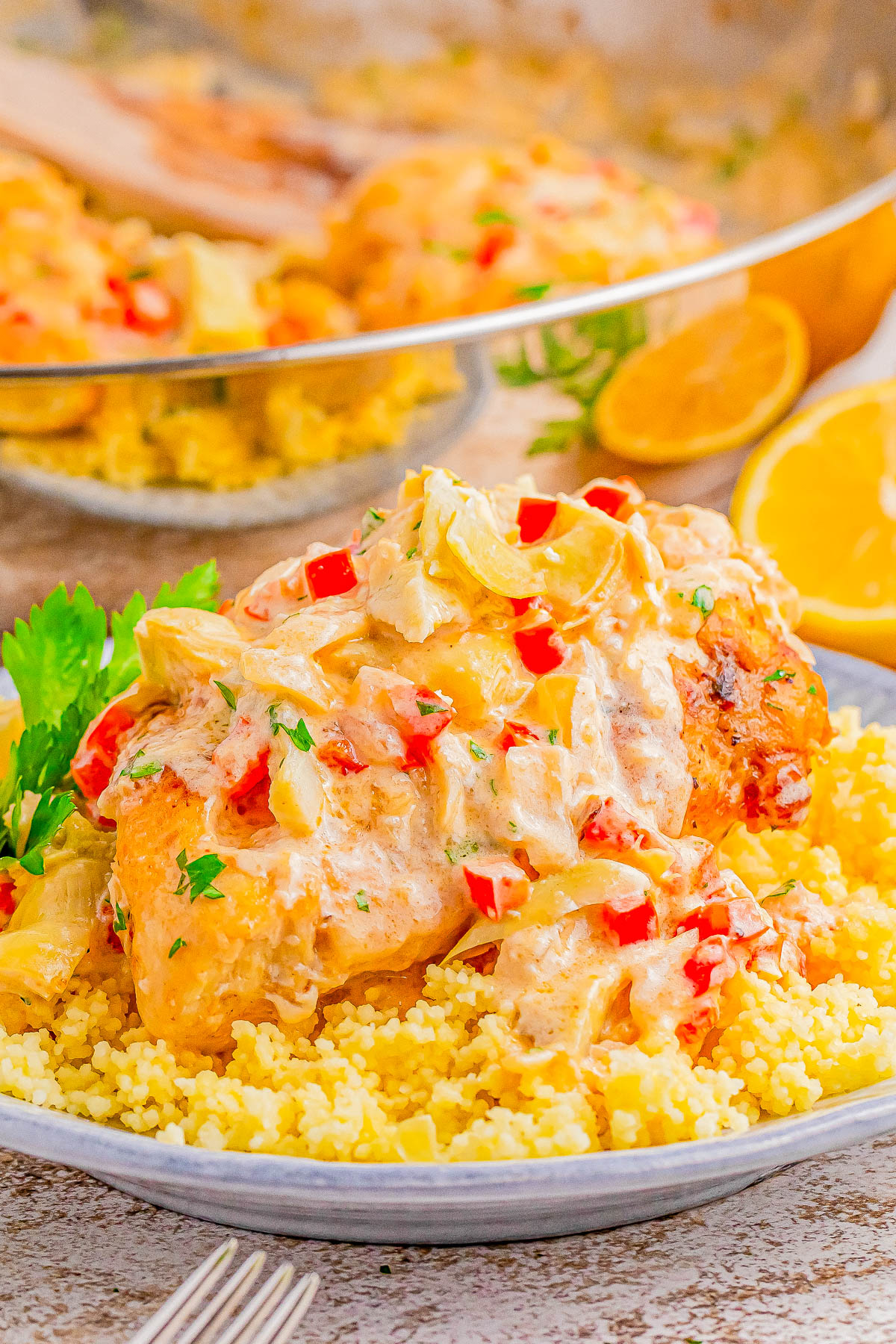 A plate with chicken topped with a creamy sauce and diced vegetables, served over a bed of couscous. There are garnishes and halved lemons in the background.