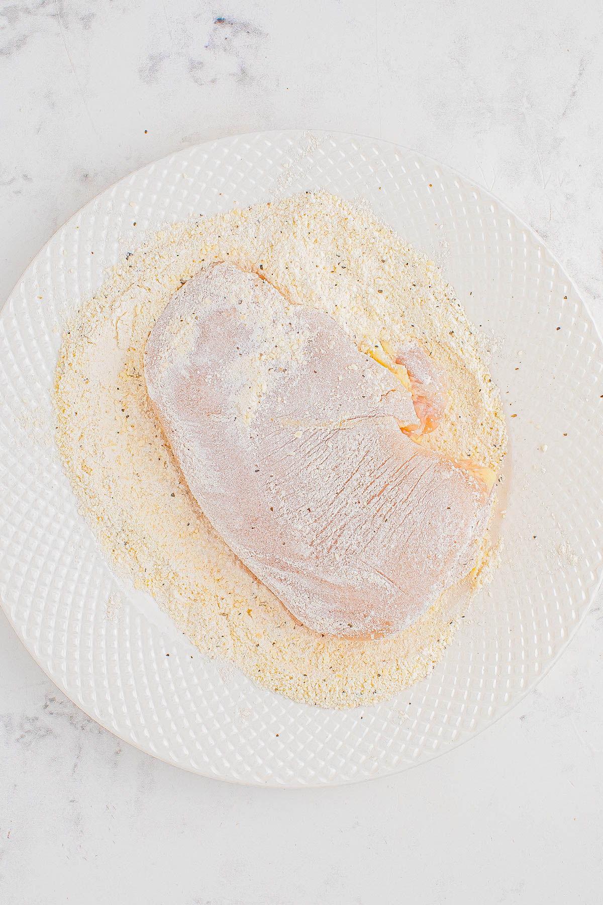 A raw piece of meat coated in flour on a white plate with a textured pattern, placed on a light-colored, marble-like surface.