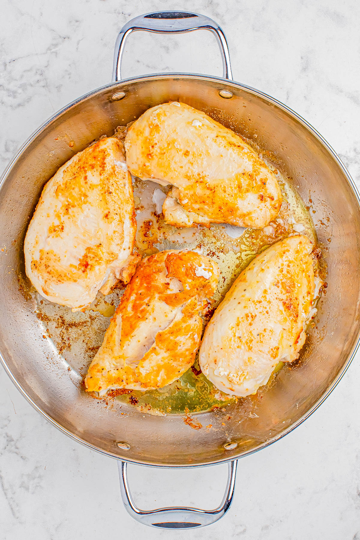 A stainless steel pan containing four cooked chicken breasts with golden-brown searing, placed on a white marble surface.